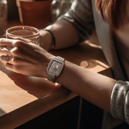 Stuhrling Original luxury rectangular silver watch with crystal bezel, Roman numerals, and stainless steel bracelet, worn on wrist while holding a glass
